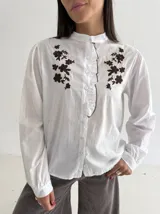 Camisa blanca de manga larga con cuello mao, cierre frontal de botones y detalle de volado. Presenta bordados florales en color marrón chocolate en la parte superior delantera.
