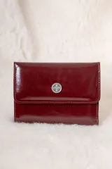 Billetera de cuero charolado color bordó con solapa frontal y aplique metálico circular con logo grabado.