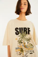 Remera extragrande de algodón color beige, de manga corta y corte holgado. Cuenta con un estampado frontal que dice 'SURF' junto a una ilustración de playa con palmeras, surfistas y la inscripción 'Rio de Janeiro'.