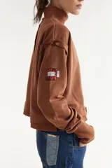 Buzo rústico marrón con cuello polera, mangas largas y calce boxy. Presenta costuras externas decorativas, puños acanalados y un bordado sutil en el pecho.