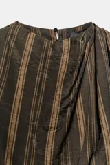 Vestido midi sin mangas de cuello redondo, con diseño de rayas verticales en tonos marrones. Presenta un detalle drapeado en el frente, volantes laterales y un bajo asimétrico. Cierre posterior con cremallera oculta.