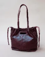 Cartera tipo tote de cuero color bordó, con diseño fruncido en los laterales y abertura central integrada como asa. Presenta una silueta flexible y espaciosa, ideal para uso diario.
