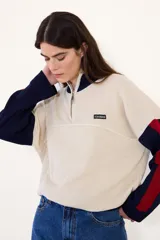 Sweater de punto acanalado con cuello alto y cierre frontal parcial. Presenta un diseño tricolor en bloques con cuerpo color crema, cuello y puños azul marino, y detalles en rojo en las mangas. Incluye una pequeña etiqueta con logo en el pecho.