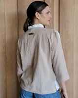 Camisa de manga tres cuartos color beige con cuello con volados y botones frontales. Presenta un detalle de color negro en la parte superior de las mangas.