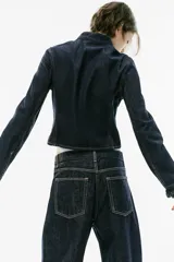 Cazadora de denim azul oscuro, de corte entallado, cuello redondo y manga larga. Cuenta con cierre frontal con alamares y costuras pespunteadas en contraste.