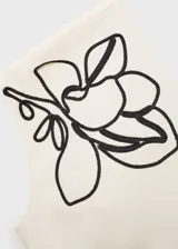 Top de algodón color crudo con diseño asimétrico de un solo hombro. Presenta un bordado lineal en color negro que representa motivos florales en el hombro y cuenta con cierre lateral oculto.