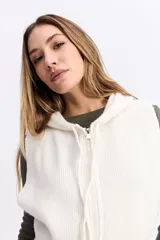 Chaleco de punto color beige, de corte corto y holgado. Presenta capucha, cierre frontal completo y terminaciones acanaladas en la cintura.