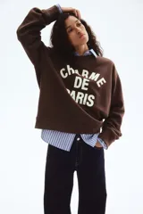 Buzo de tejido de punto color marrón oscuro, con cuello redondo, hombros caídos y puños y bajo acanalados. Presenta un estampado frontal en color crema con el texto "CHARME DE PARIS 1998".