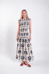 Vestido largo sin mangas de color beige con estampado floral y vegetal en color azul marino. Cuenta con un cuerpo ajustado mediante fruncidos elásticos (smock) y falda larga con vuelo y corte en capas.