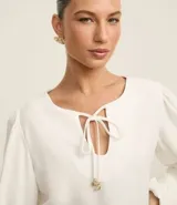 Blusa blanca de poliéster satinado, con mangas largas abullonadas, cuello con abertura en forma de gota y lazo con colgantes de corazón.