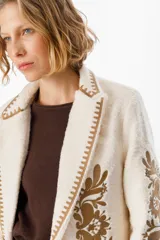 Blazer de textura suave en color crudo, con solapas clásicas y bordados florales en tonos tierra en el frente y las mangas. Presenta detalles de costura decorativa en los bordes de las solapas.