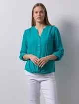 Blusa blanca de gasa marca Zac & Rachel, con cuello a la base, escote en V, abertura central con botones, bolsillo aplicado en el pecho y mangas largas regulables con presilla y botón. Tiene terminación opcional de nudo en el ruedo.