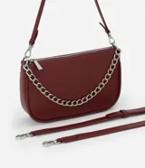 Cartera tipo baguette color bordó, confeccionada en poliuretano con acabado liso. Presenta una cadena decorativa plateada en el frente, cierre superior con cremallera y correa larga ajustable y desmontable.