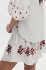 Blusa blanca de gasa de algodón con mangas 3/4 abullonadas y escote en V con botones. Cuenta con bordados en tonos marrones y naranjas en la zona frontal y las mangas.