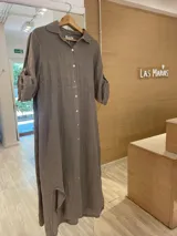 Vestido camisero largo de lino, color gris topo, con cuello camisero, manga larga enrollada y cierre frontal de botones de nácar. Presenta un corte en la cintura y abertura lateral en el bajo.
