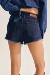 Short de denim azul oscuro con tiro bajo y ruedo deshilachado. Cuenta con bolsillos delanteros, cierre frontal con botón metálico personalizado y costuras en contraste.