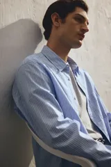 Camisa celeste y blanca a rayas verticales, de corte holgado, manga larga, cuello inglés, bolsillo en el pecho y cierre con botones.
