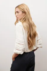 Camisa de color crudo con diseño romántico, cuello redondo con cordón ajustable terminado en borlas, y mangas tres cuartos abullonadas. Presenta detalles de bordados calados verticales en el frente y horizontales en las mangas.