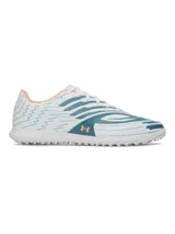 Championes de fútbol Under Armour Shadow Select 4 Turf, diseñados para césped artificial. Presentan un diseño aerodinámico en color blanco con detalles en azul petróleo y acentos en naranja. Cuentan con empeine sintético texturizado para mejor control, mediasuela Charged Cushioning para amortiguación reactiva y suela de goma con tacos multidireccionales.