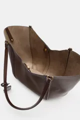 Bolso tipo shopper de hombro en color beige, con diseño estructurado, doble asa y detalle de pespuntes verticales en el cuerpo. Incluye un colgante decorativo tipo etiqueta y cierre imantado.