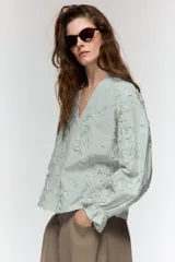 Blusa de escote pico y manga larga con puño elástico y volante. Presenta un diseño de rayas verticales finas en azul claro y blanco, con flores en relieve.
