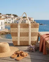 Bolso tipo tote confeccionado en paja tejida con diseño de rayas horizontales en tonos beige y marrón. Cuenta con doble asa de hombro de lona clara y cierre superior.