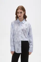 Camisa de algodón con diseño de rayas verticales finas en color celeste y blanco, con apliques florales bordados en relieve. Presenta cuello camisero, cierre frontal con botones y mangas largas con puños abotonados.