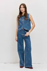 Pantalón de jean azul de tiro medio y corte wide leg, con pequeños bordados de motivos paisley en hilo marrón distribuidos por toda la prenda. Cuenta con bolsillos clásicos, presillas para cinto y cierre frontal con botón y cremallera.