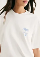 Remera blanca de manga corta, corte oversized, cuello redondo y pequeño estampado azul en el pecho con diseño de palmera y texto.
