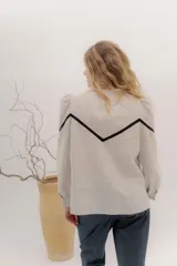 Camisa de lino color beige con mangas abullonadas y detalles de puntilla negra en forma de V en el frente. Presenta cuello a la base con borde desflecado, cierre frontal con botones forrados y puños con alforzas.