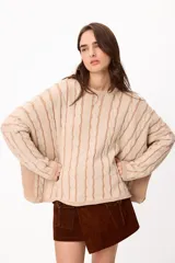 Sweater de punto con diseño de trenzas verticales, cuello redondo y corte crop. Presenta una combinación de colores crudo y camel, con mangas largas y ajuste holgado.
