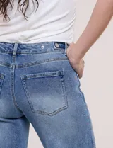 Jean wide leg de tiro medio, color azul claro, confeccionado en denim de algodón con un toque de elastano. Cuenta con cinco bolsillos, presillas para cinturón y cierre frontal con botón. El diseño destaca por su pretina regulable con cruce y broches metálicos.