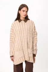 Sweater de punto con diseño de trenzas verticales, color crudo y camel, con bolsillos frontales y detalle de logo bordado. Presenta un corte holgado y mangas largas.