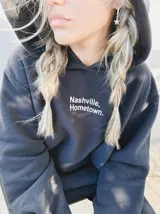 Buzo con capucha de corte amplio y textura suave, color negro, con estampa frontal en color blanco que dice 'Nashville, Hometown'.