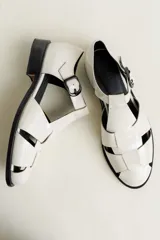 Sandalia cerrada tipo pescador, color blanco o hueso brillante, con tiras entrecruzadas en el empeine y cierre de hebilla ajustable en el lateral. Posee suela y taco bajo de color negro.