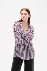 Camisa de corte holgado con estampado de rayas verticales en tonos bordó y blanco. Presenta cuello clásico, cierre frontal con botones, mangas largas con puños abotonados y un bolsillo frontal en el pecho.