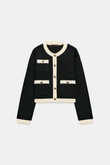 Chaqueta de punto de corte recto y manga larga, con cuello redondo. Presenta un diseño bicolor en negro y crema, con cierre frontal de botones dorados y bolsillos de plastrón en el delantero con detalles en contraste.