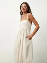 Vestido midi color beige claro, de corte amplio y fluido, con tirantes finos ajustables mediante lazos en los hombros, escote recto y prácticos bolsillos laterales.