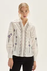 Blusa blanca de algodón con bordados geométricos en tonos negros y lilas, cuello clásico, cierre frontal con botones y mangas largas abullonadas con puños. Presenta detalles de vainillas y calados en el frente y mangas.