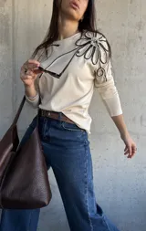 Remera manga larga de color beige, con detalle de aplique floral tridimensional en el hombro confeccionado con cordones en blanco y negro.