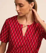 Blusa de crepé color rojo con estampado geométrico de rombos en negro y blanco. Tiene cuello redondo con abertura en forma de gota y detalle metálico, mangas cortas y corte recto.