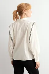 Camisa de color blanco con cuello clásico, cierre frontal de botones y mangas largas con puños. Presenta detalles de bordado geométrico en contraste a lo largo de los hombros y la parte superior de la espalda.