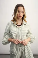 Camisa a rayas verticales color beige y blanco, con cuello clásico, cierre frontal con botón dorado, mangas largas abullonadas con puños elásticos, dos bolsillos frontales y corte holgado.