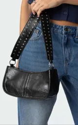 Cartera tipo baguette de cuero sintético negro con acabado brillante, cierre superior y correa ancha decorada con tachas metálicas plateadas.