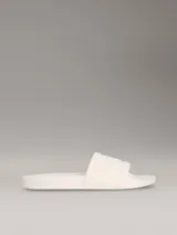 Ojotas tipo slide de color blanco hueso, con tira ancha acolchada y monograma de CK en relieve. La suela es de espuma.