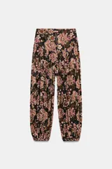 Pantalón bombacho de tiro alto con estampado floral en tonos rosados y verdes sobre fondo negro. Presenta un detalle de hilo metalizado brillante y bajo acabado abullonado con elástico.