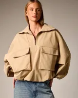 Chaqueta color beige de estilo utilitario con cuello alto y cierre frontal parcial. Presenta mangas amplias con puños elásticos y dos bolsillos frontales tipo cargo con solapa.