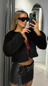 Chaqueta corta de piel sintética en color negro, con diseño de cuello redondo y mangas largas. Presenta una textura suave y voluminosa, con un corte recto y acabado elegante.