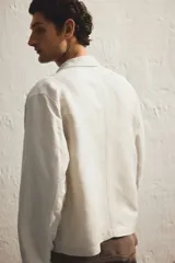 Campera color crema confeccionada en mezcla de lino y viscosa, con cuello clásico, cierre frontal, bolsillos laterales insertados al bies con botón a presión oculto, costura decorativa en la espalda y botón a presión en los puños.