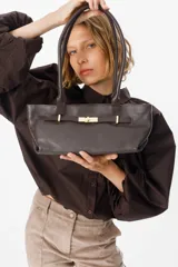 Cartera de mano tipo baguette confeccionada en cuero vacuno con textura granulada. Presenta un diseño rectangular con doble asa fija y un cierre frontal metálico en tono dorado tipo torniquete.